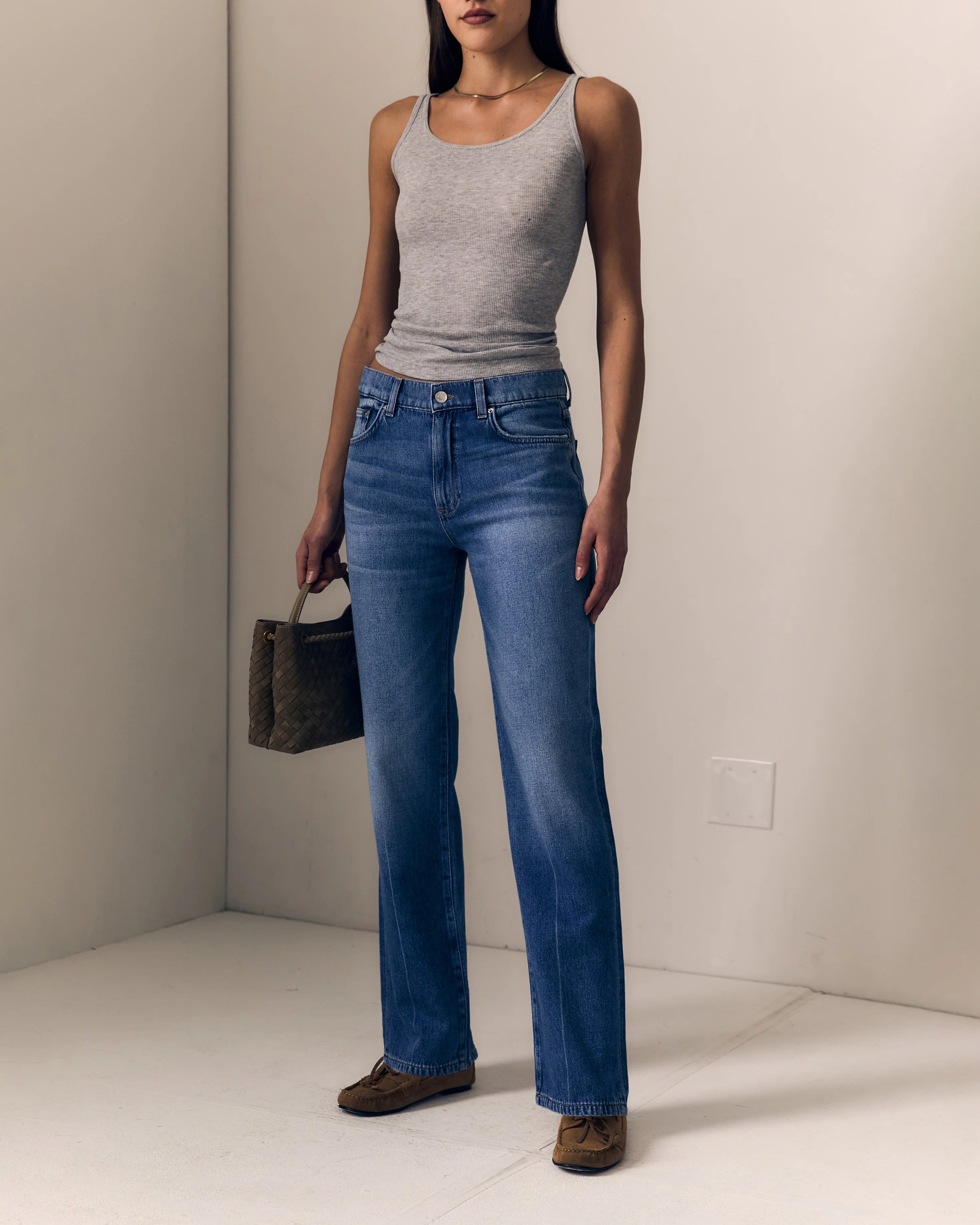 A model wearing a pair of high-waisted, straight-leg blue jeans paired with a light gray ribbed tank top and brown suede loafers, holding a dark patterned handbag in a minimalist studio setting.

Le Jean Mid 90's Straight in North Star. Available at Edit by Elaine Turner in Houston, TX. Full model front view. 