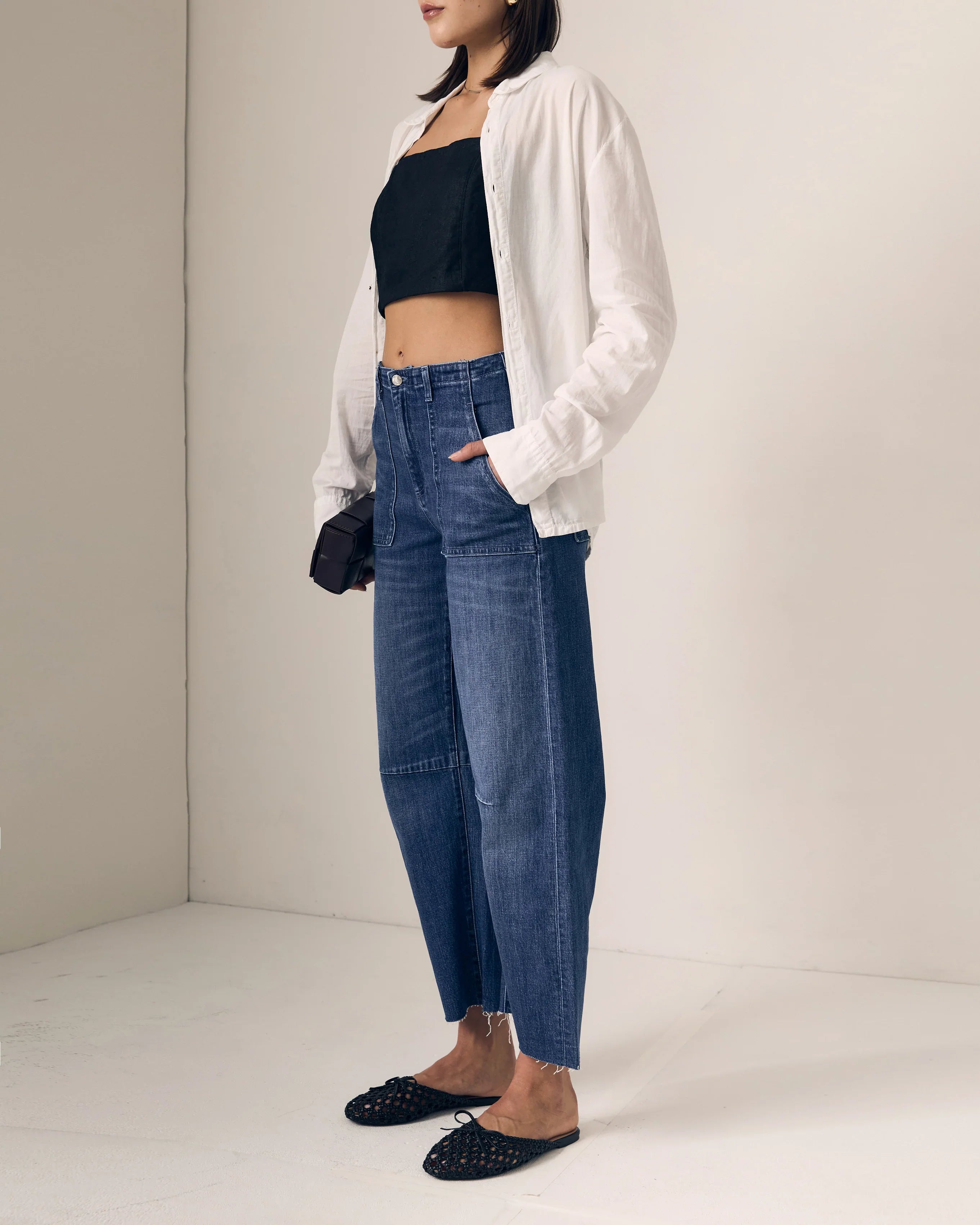 A model wearing a pair of relaxed, wide straight leg blue jeans with a black crop top, white button-up shirt, and black studded flats, standing in a minimalist studio setting.

Le Jean Utility Soft Barrel in London Calling. Available at Edit by Elaine Turner in Houston, TX. Full model side view. 