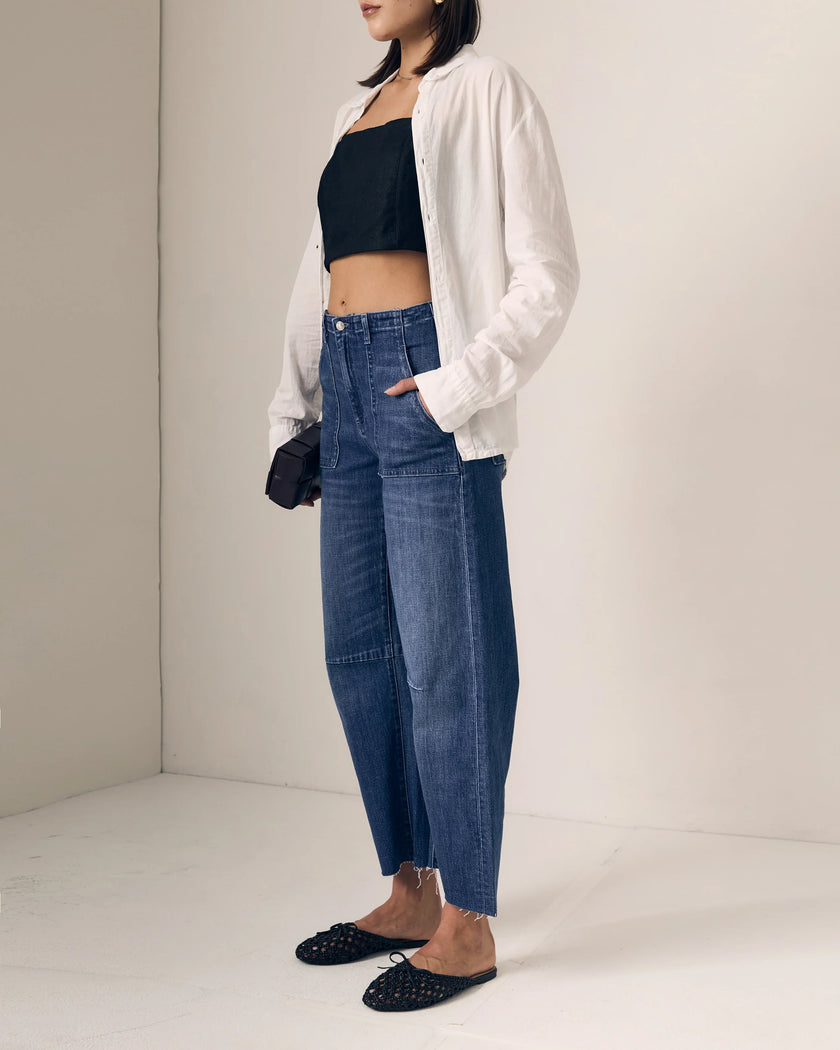 A model wearing a pair of relaxed, wide straight leg blue jeans with a black crop top, white button-up shirt, and black studded flats, standing in a minimalist studio setting.

Le Jean Utility Soft Barrel in London Calling. Available at Edit by Elaine Turner in Houston, TX. Full model side view. 