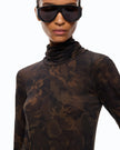 A close-up of a model wearing a sheer, long-sleeve turtleneck top with a dark brown and black floral camouflage pattern, paired with oversized black sunglasses, against a plain white background.

PINKO- Turtleneck Sweater with Floral Camouflage Print in Green & Black Multicolor. Available at Edit by Elaine Turner in Houston, TX. Close up model view. 