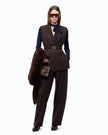 A model stands against a white studio background wearing a dark brown tailored vest and matching trousers over a navy long-sleeve top, accessorized with a navy neck bow, dark sunglasses, a dark brown fur-look coat draped over one arm, and a small dark brown handbag.

PIINKO- Vest with Open Sides and Belt in Beyond the Dark Brown. Available at Edit by Elaine Turner in Houston, TX. Full Model Front View. 