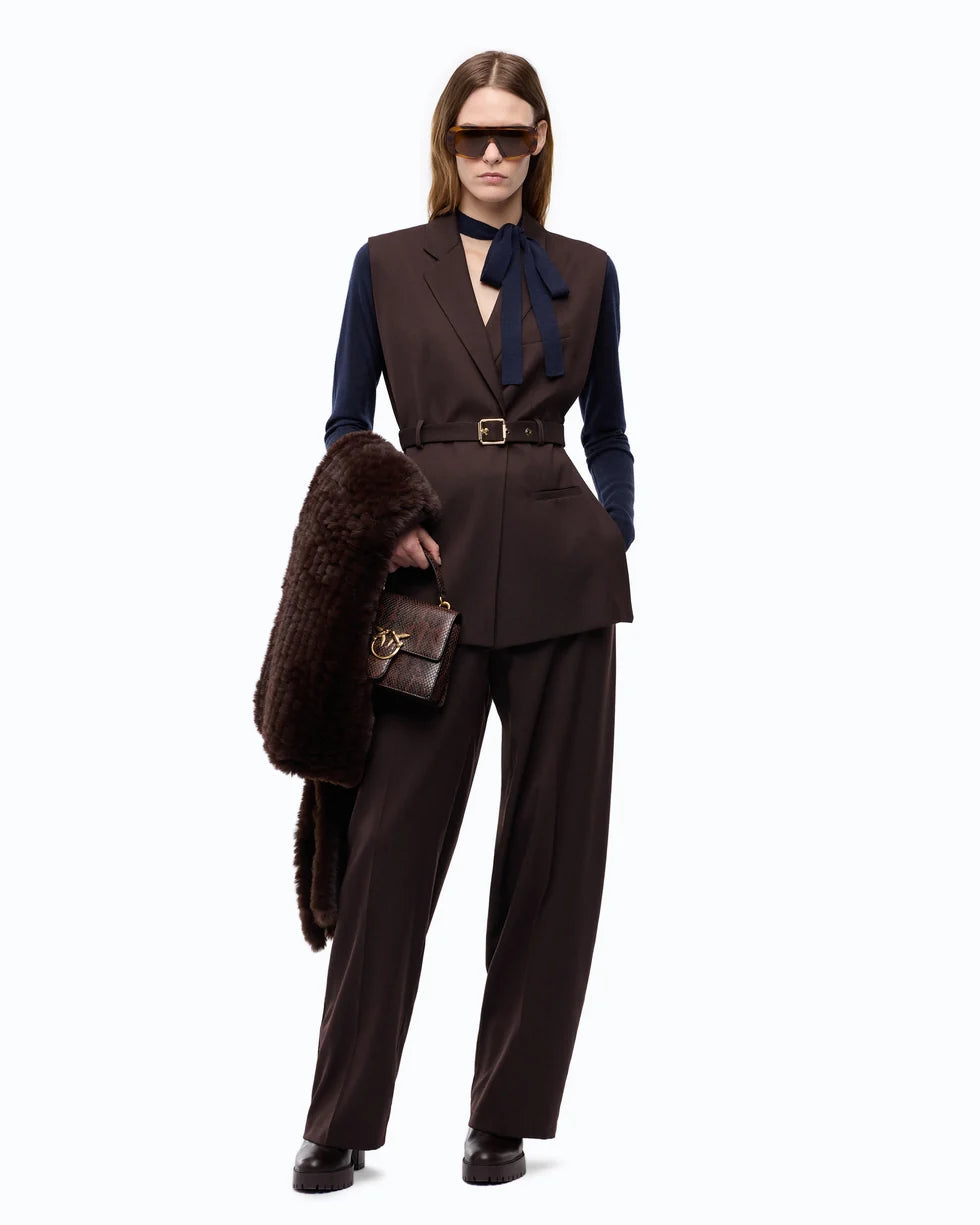 A model stands against a white studio background wearing a dark brown tailored vest and matching trousers over a navy long-sleeve top, accessorized with a navy neck bow, dark sunglasses, a dark brown fur-look coat draped over one arm, and a small dark brown handbag.

PIINKO- Vest with Open Sides and Belt in Beyond the Dark Brown. Available at Edit by Elaine Turner in Houston, TX. Full Model Front View. 