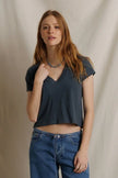 A woman with long light brown hair wearing a dark slate blue-gray short-sleeve V-neck crop top and blue jeans, standing against a draped beige fabric backdrop.

Perfect White Tee - Alanis in Turbulence. Available at Edit by Elaine Turner in Houston, TX. Model front view. 