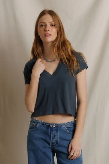 A woman with long light brown hair wearing a dark slate blue-gray short-sleeve V-neck crop top and blue jeans, standing against a draped beige fabric backdrop.

Perfect White Tee - Alanis in Turbulence. Available at Edit by Elaine Turner in Houston, TX. Model front view. 
