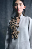 A woman wearing a light gray knit cardigan with a large, textured brown knitted floral and leaf embellishment on the chest, set against a dark gray studio background.

Psophia - long sleeve knit cardigan with floral applique. Available at Edit by Elaine Turner in Houston, TX. Close - up model view. 