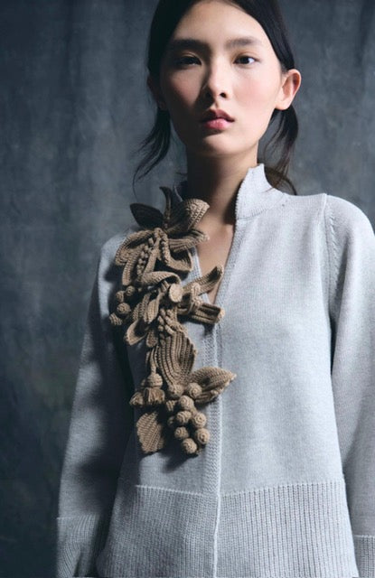 A woman wearing a light gray knit cardigan with a large, textured brown knitted floral and leaf embellishment on the chest, set against a dark gray studio background.

Psophia - long sleeve knit cardigan with floral applique. Available at Edit by Elaine Turner in Houston, TX. Close - up model view. 