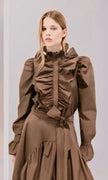 A model wearing a brown long-sleeve blouse with a high ruffled collar, gathered waist, and matching tie belt, set against a plain light gray background.

Psophia long sleeve taffeta blouse in truffle. Available at Edit by Elaine Turner in Houston, TX. Model front view. 