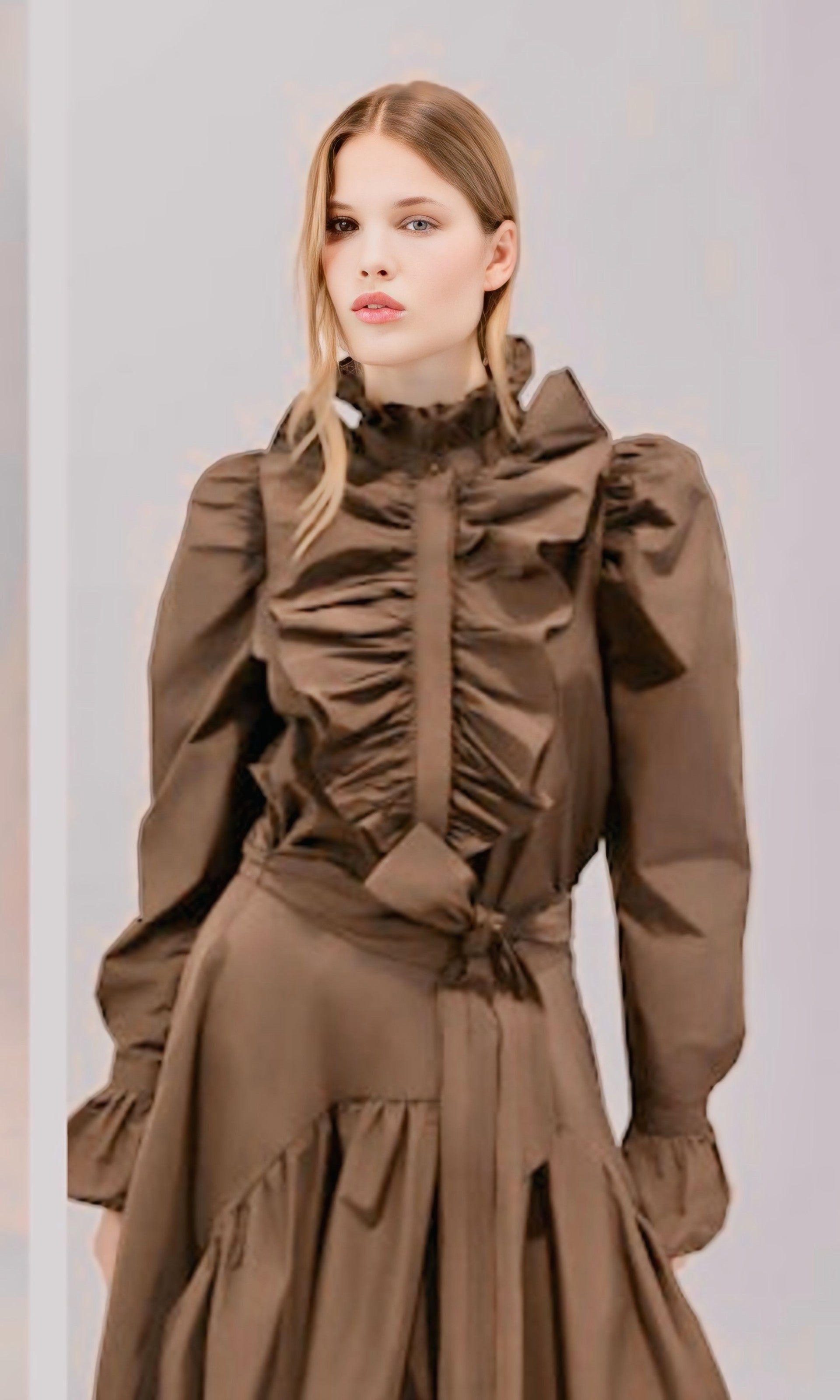 A model wearing a brown long-sleeve blouse with a high ruffled collar, gathered waist, and matching tie belt, set against a plain light gray background.

Psophia long sleeve taffeta blouse in truffle. Available at Edit by Elaine Turner in Houston, TX. Model front view. 