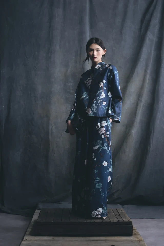 A model stands on a wooden platform wearing a dark navy blue kimono-style outfit with a white and pink floral print, set against a dark gray fabric backdrop.

Psophia Palazzo Trousers in Almond Blossom. Available at Edit by Elaine Turner in Houston, TX. Full Model View. 