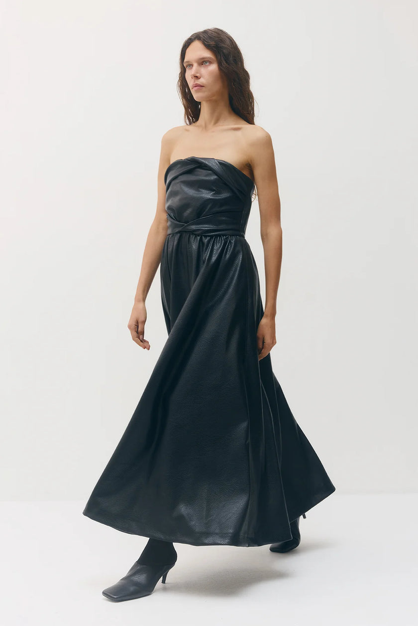 A woman modeling a strapless black faux-leather maxi dress with a gathered bodice and flowing skirt, paired with black pointed-toe heeled boots, against a plain light gray studio background.

Sabina Musayev Garner Dress in black. Available at Edit by Elaine Turner in Houston, TX. Side Model View. 