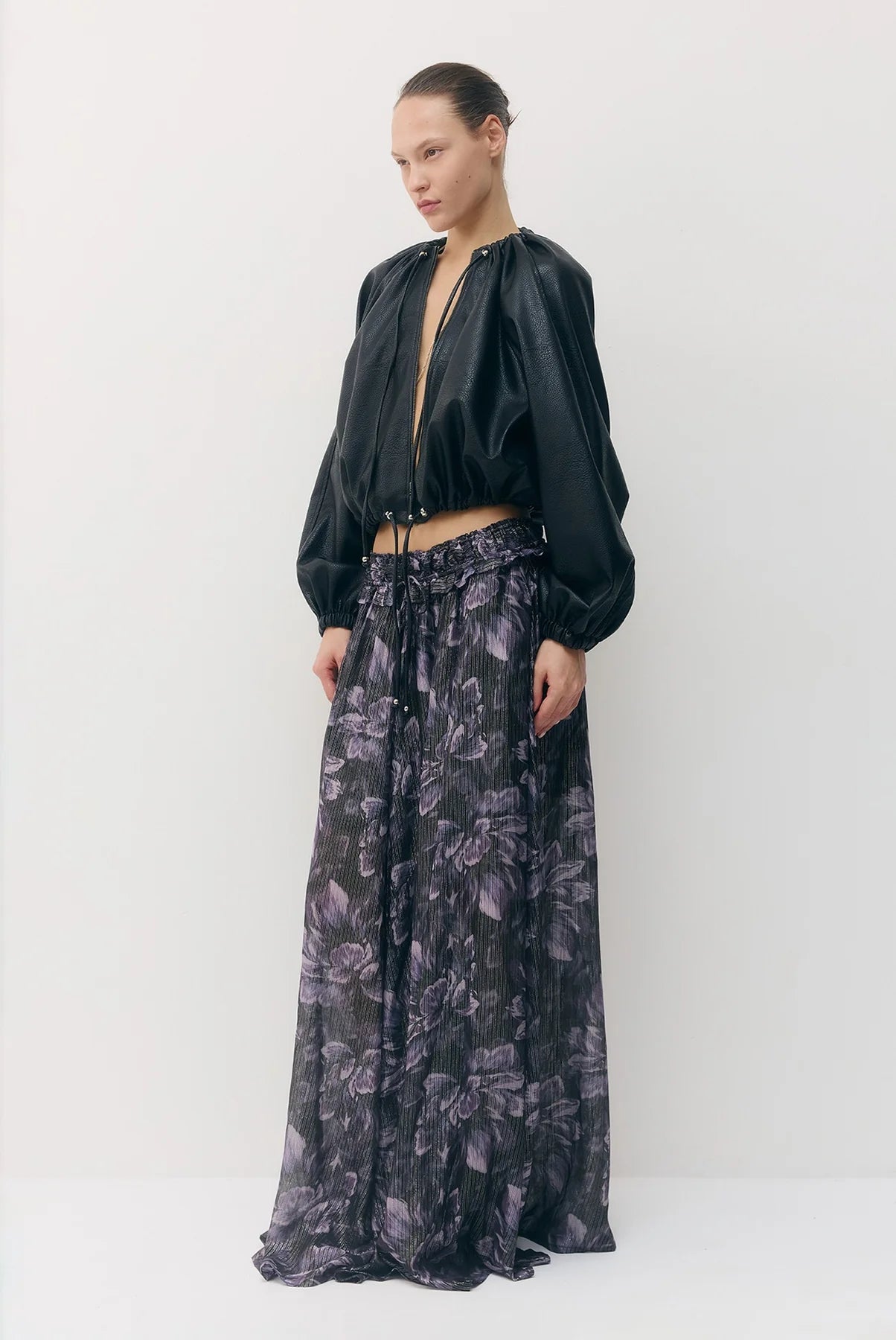 A model stands against a plain light gray studio background wearing a black vegan leather cropped jacket with long sleeves and a dark floral maxi skirt with purple and gray tones.

Sabina Musayev Tomas Jacket in black. Available at Edit by Elaine Turner in Houston, TX. Front Side Model View. 