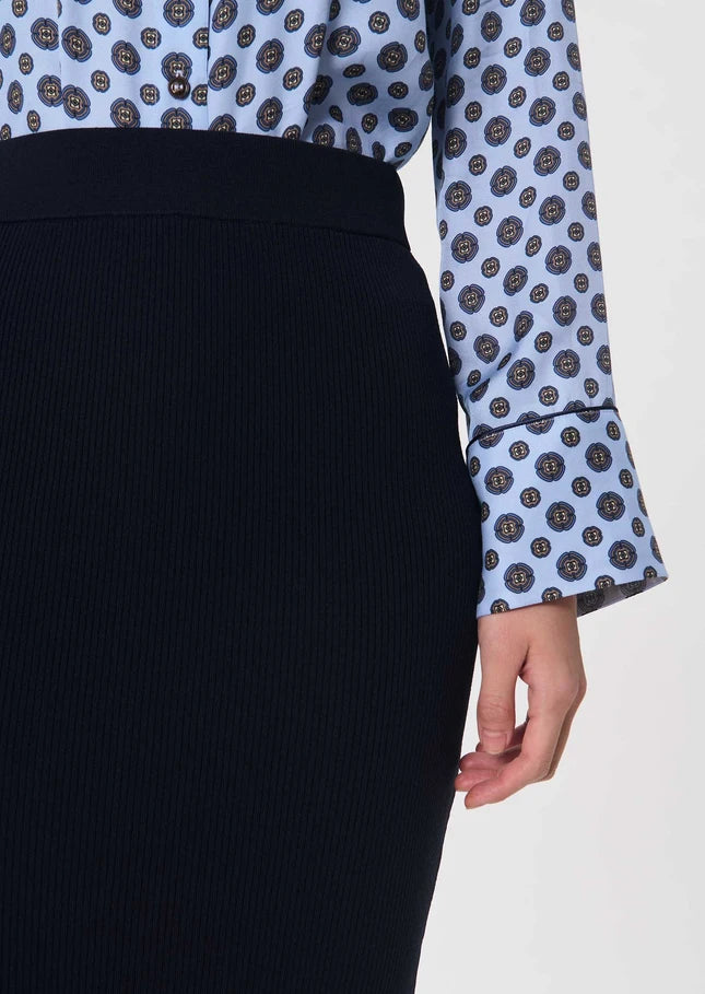 A close-up of a person wearing a light blue long-sleeve blouse with a dark circular pattern and a dark navy ribbed skirt, shown against a plain light background.

Tara Jarmon- Jeannette Knit Skirt in Nuit. Available at Edit by Elaine Turner in Houston, TX. Model Close Up View. 