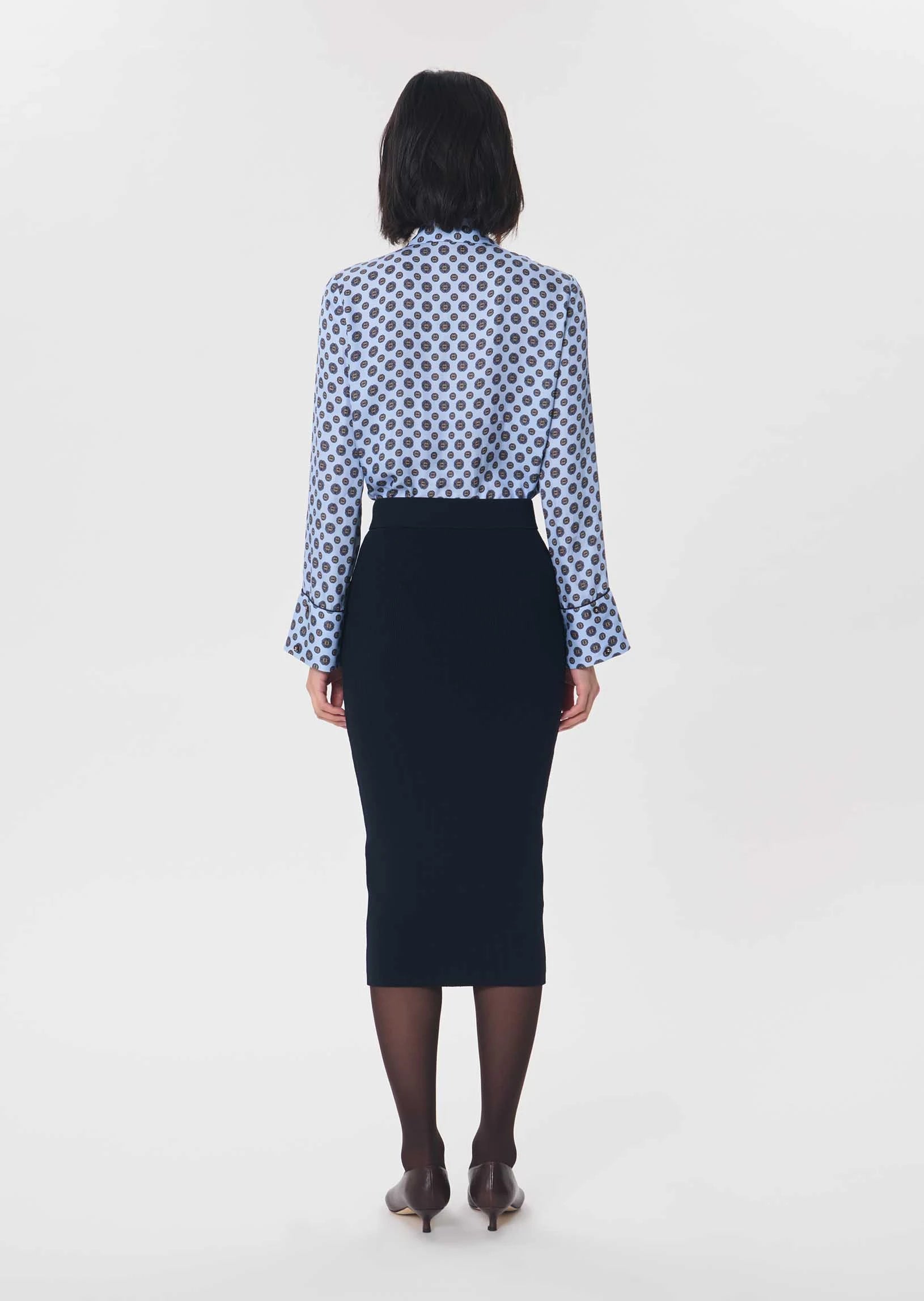 A woman viewed from the back wearing a long-sleeve button-up shirt with a blue and white geometric pattern and a dark navy pencil skirt, paired with sheer black tights and brown heels, against a plain light gray studio background.

Tara Jarmon- Jeannette Knit Skirt in Nuit. Available at Edit by Elaine Turner in Houston, TX. Back Model View. 
