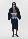 A model stands against a light gray studio background wearing a dark navy blue jacket and matching skirt set, layered over a gray knit sweater and a blue-and-white patterned collared shirt, with dark brown pointed-toe heels and dark sunglasses.

Tara Jarmon- Jeannette Knit Skirt in Nuit. Available at Edit by Elaine Turner in Houston, TX. Front Model View. 