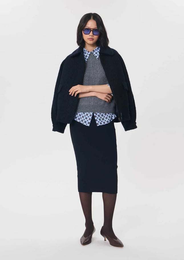 A model stands against a light gray studio background wearing a dark navy blue jacket and matching skirt set, layered over a gray knit sweater and a blue-and-white patterned collared shirt, with dark brown pointed-toe heels and dark sunglasses.

Tara Jarmon- Jeannette Knit Skirt in Nuit. Available at Edit by Elaine Turner in Houston, TX. Front Model View. 