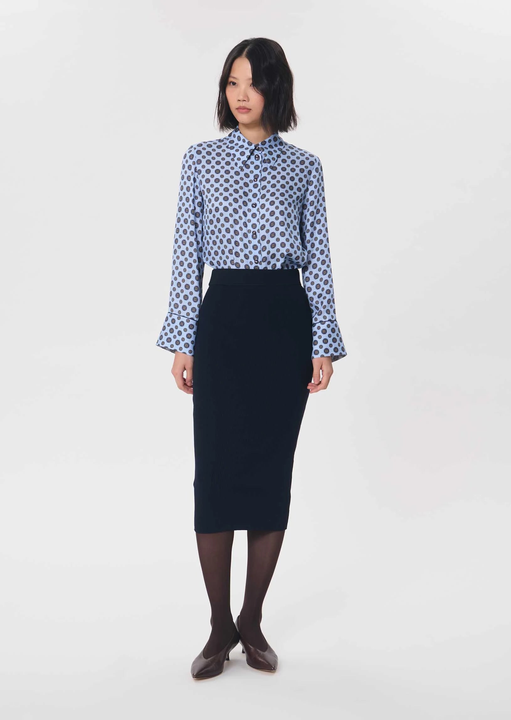 A woman stands against a plain light gray studio background wearing a long-sleeve button-up shirt with a blue and white geometric pattern, paired with a dark navy pencil skirt, dark tights, and dark brown shoes.

Tara Jarmon- Jeannette Knit Skirt in Nuit. Available at Edit by Elaine Turner in Houston, TX. Front Model View. 