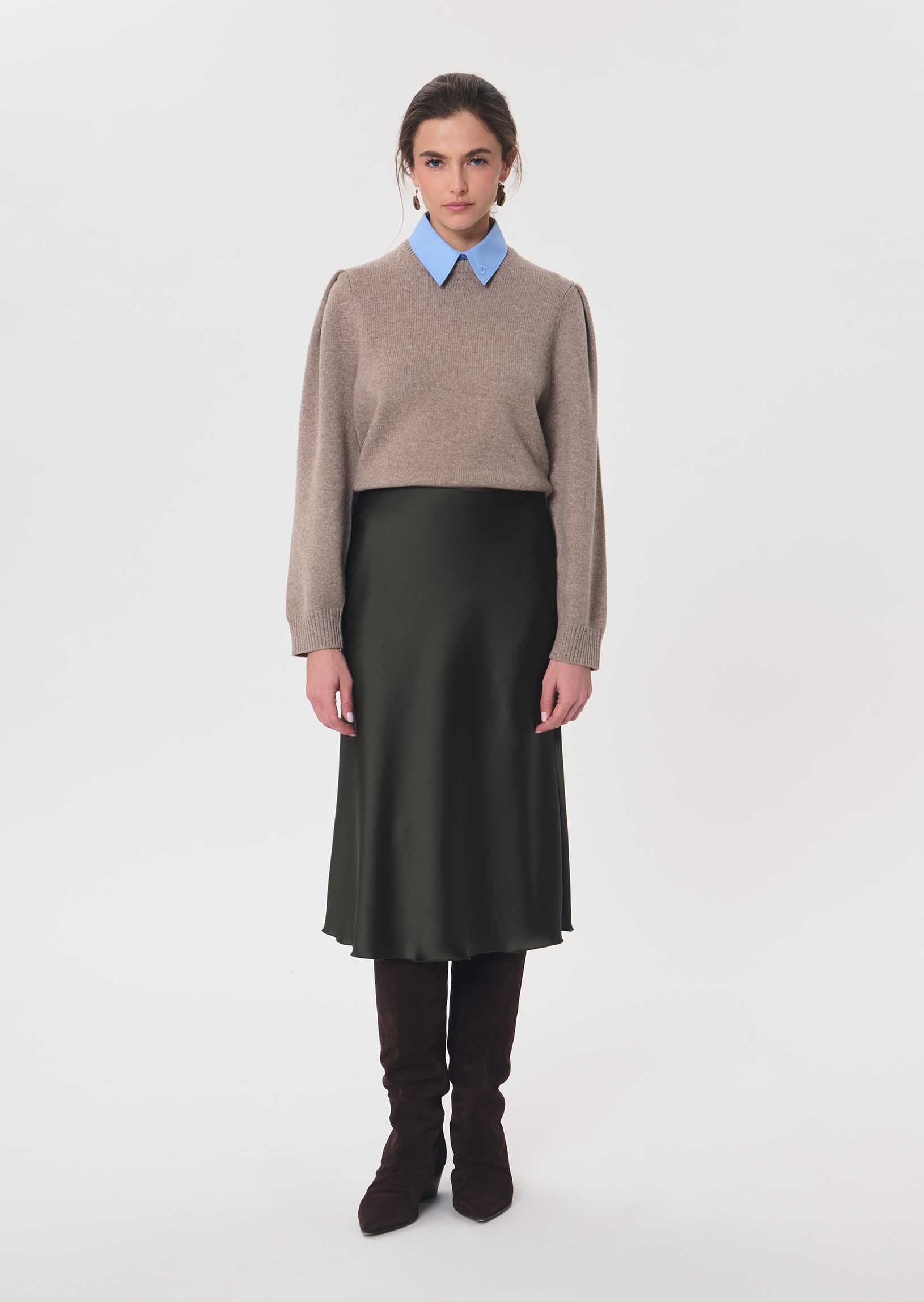 A model stands against a light gray studio background wearing a taupe knit sweater over a light blue collared shirt, paired with a dark olive green midi skirt, dark brown tights, and dark brown knee-high boots.

Tara Jarmon- Julia Skirt in Kaki. Available at Edit by Elaine Turner in Houston, TX. Model front view. 